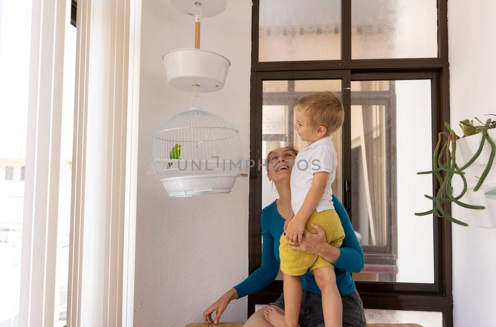 Smiling mother lifting her toddler son to see a parakeet in a white cage hanging on the balcony