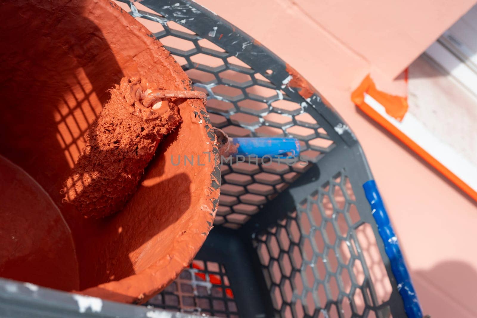 A paint roller rests in a bucket filled with reddish-brown paint. Surrounding tools and a work environment indicate ongoing home improvement activities in good weather.