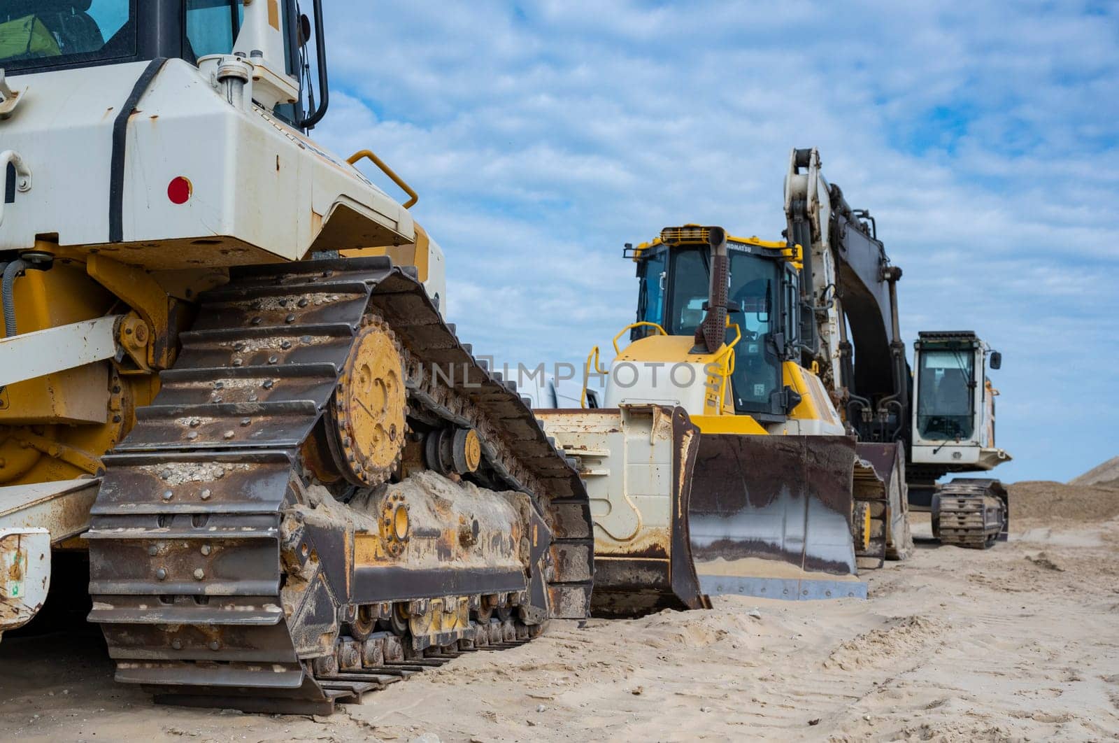 Heavy machinery is positioned along the sandy coastline, actively involved in erosion control and coastal protection efforts under a cloudy sky.