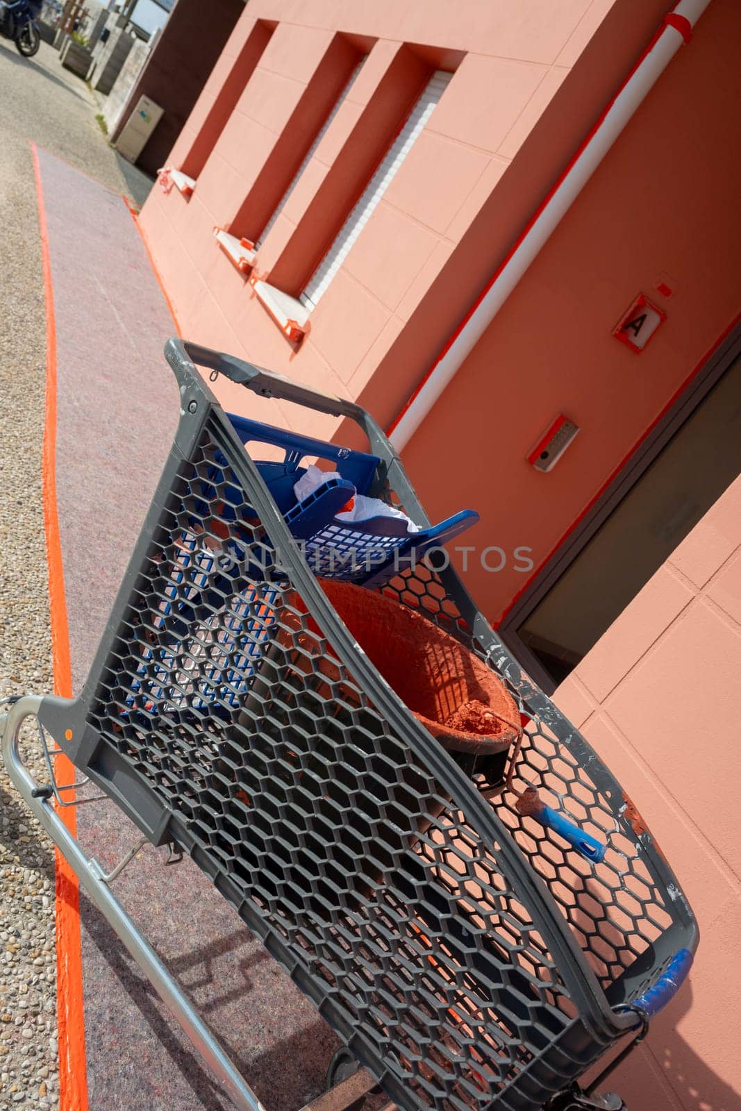 A paint roller rests in a bucket filled with reddish-brown paint. Surrounding tools and a work environment indicate ongoing home improvement activities in good weather.
