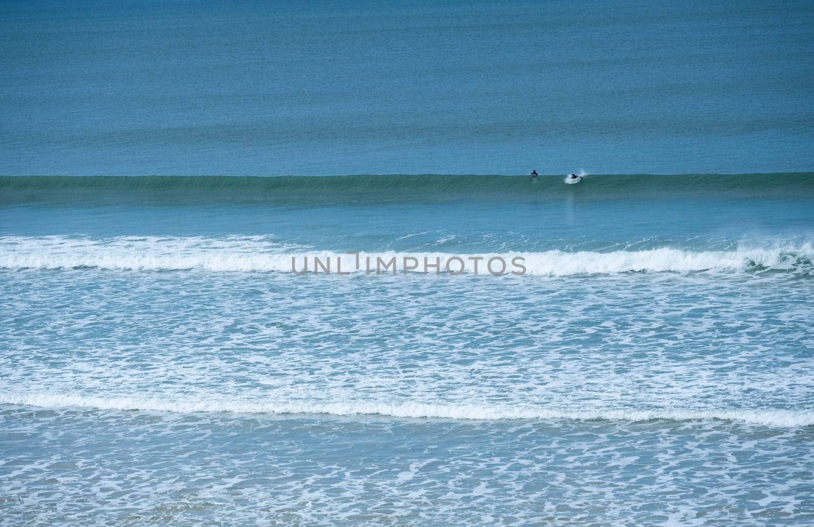 Two surfers navigate the rolling waves at Biscarrosse Beach during a serene day. The calm waters reflect the azure sky, creating a perfect surfing environment.