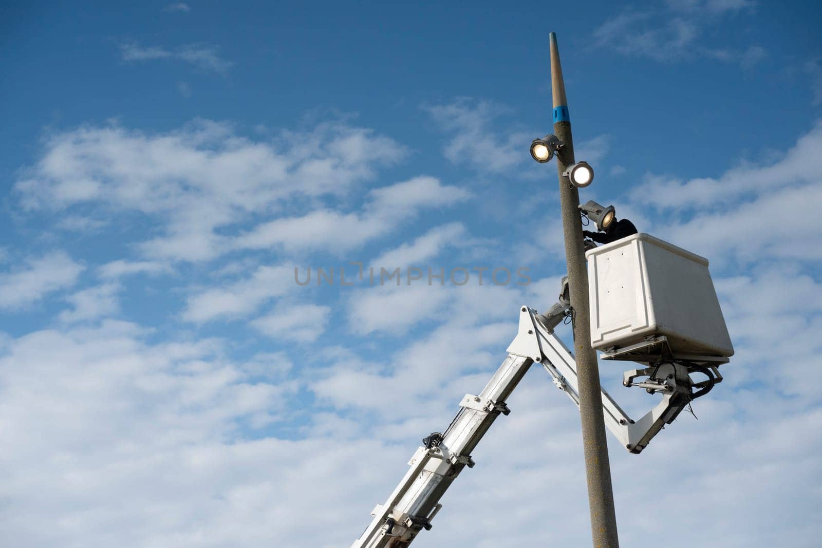 A worker is performing maintenance on a light pole, utilizing a lift for safety and access. The backdrop features a vibrant blue sky, dotted with wispy clouds.