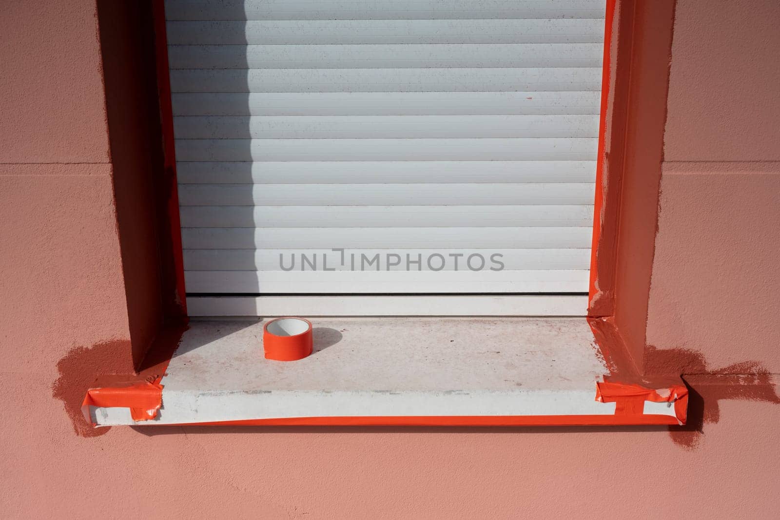 A window with white shutters is being painted with bright orange trim, showcasing fresh paint on the ledge and masking tape neatly applied around the edges for a clean finish.