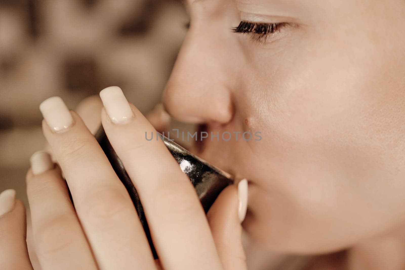 Close-up of a woman with light skin and neutral-toned nails delicately sipping from a small, dark-colored cup. Her expression is serene.