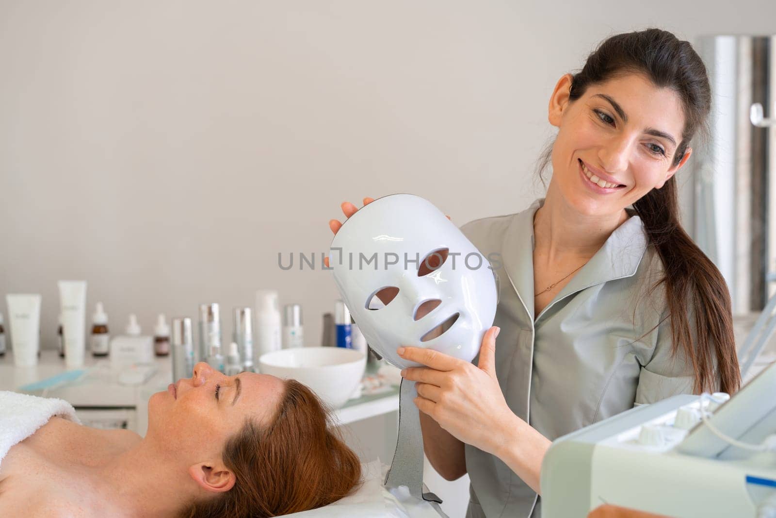 Happy female cosmetologist holds modern hydrafacial mask next to her client in salon, preparing for rejuvenating skincare treatment while creating calm and professional atmosphere
