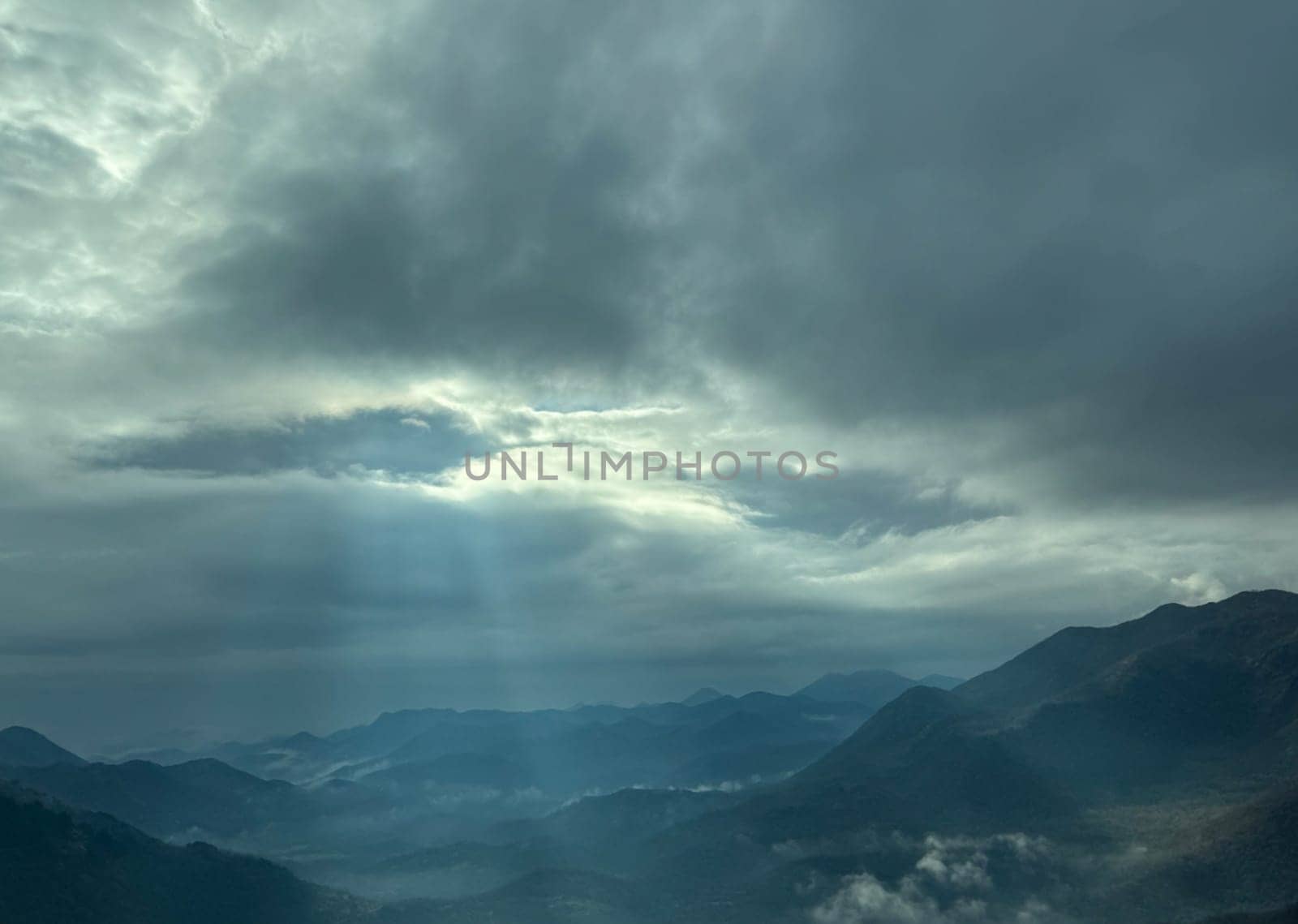 A dramatic sky with thick clouds allowing beams of sunlight to stream through, illuminating the misty mountain landscape below.