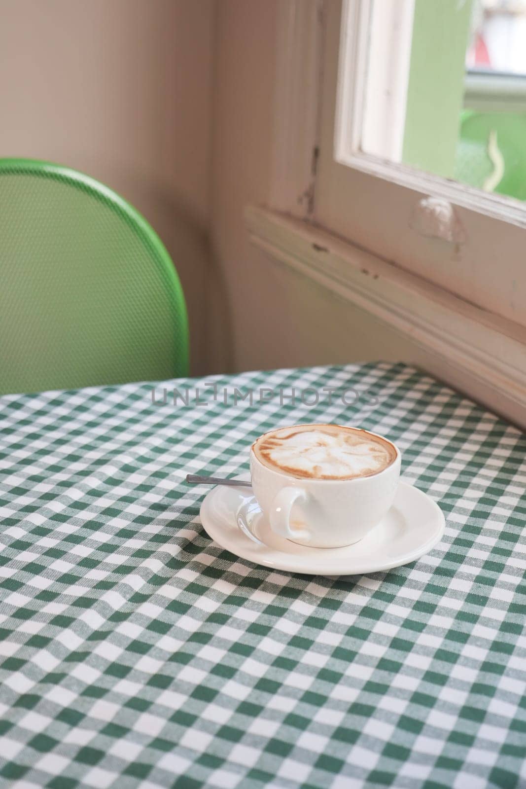 A steaming cup of coffee sits on a table with checkered cloth near a window.