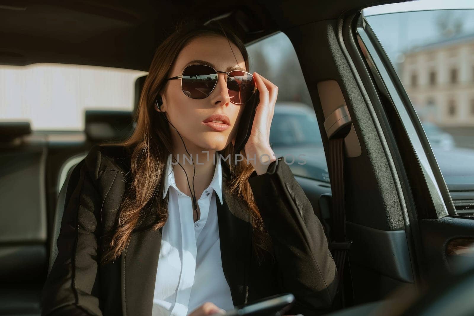 A stylish woman in sunglasses adjusts her hair while sitting in the backseat of a car, bathed in warm sunlight.