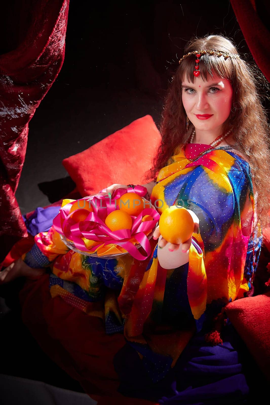 Vibrant sari-clad woman poses with a basket of oranges, showcasing her elegance in a colorful Indian-inspired setting.