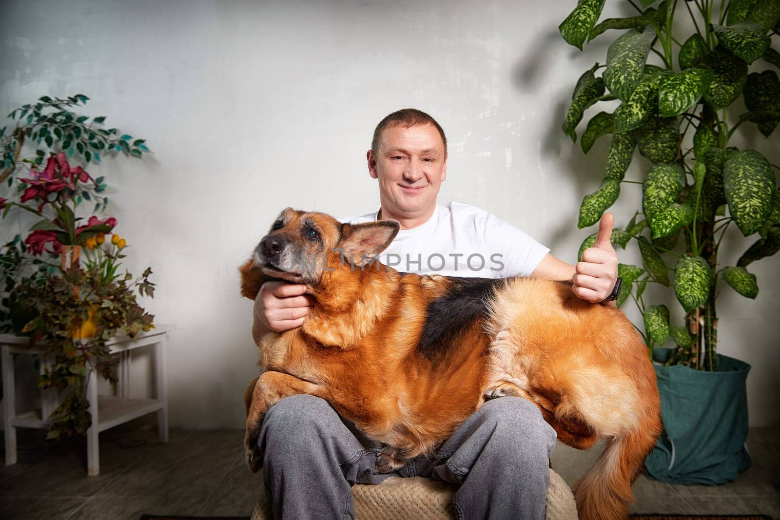 A man sits comfortably with his German Shepherd, smiling and giving a thumbs up while lounging indoors among plants.