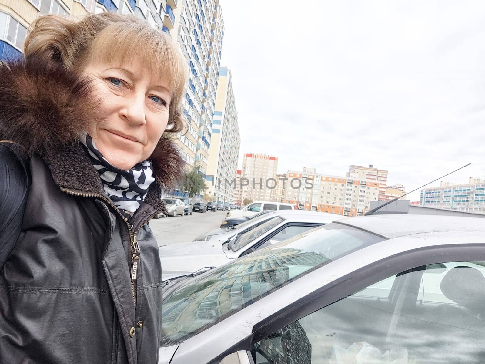 Woman smiles while posing next to her vehicle in a busy city parking area during overcast weather.
