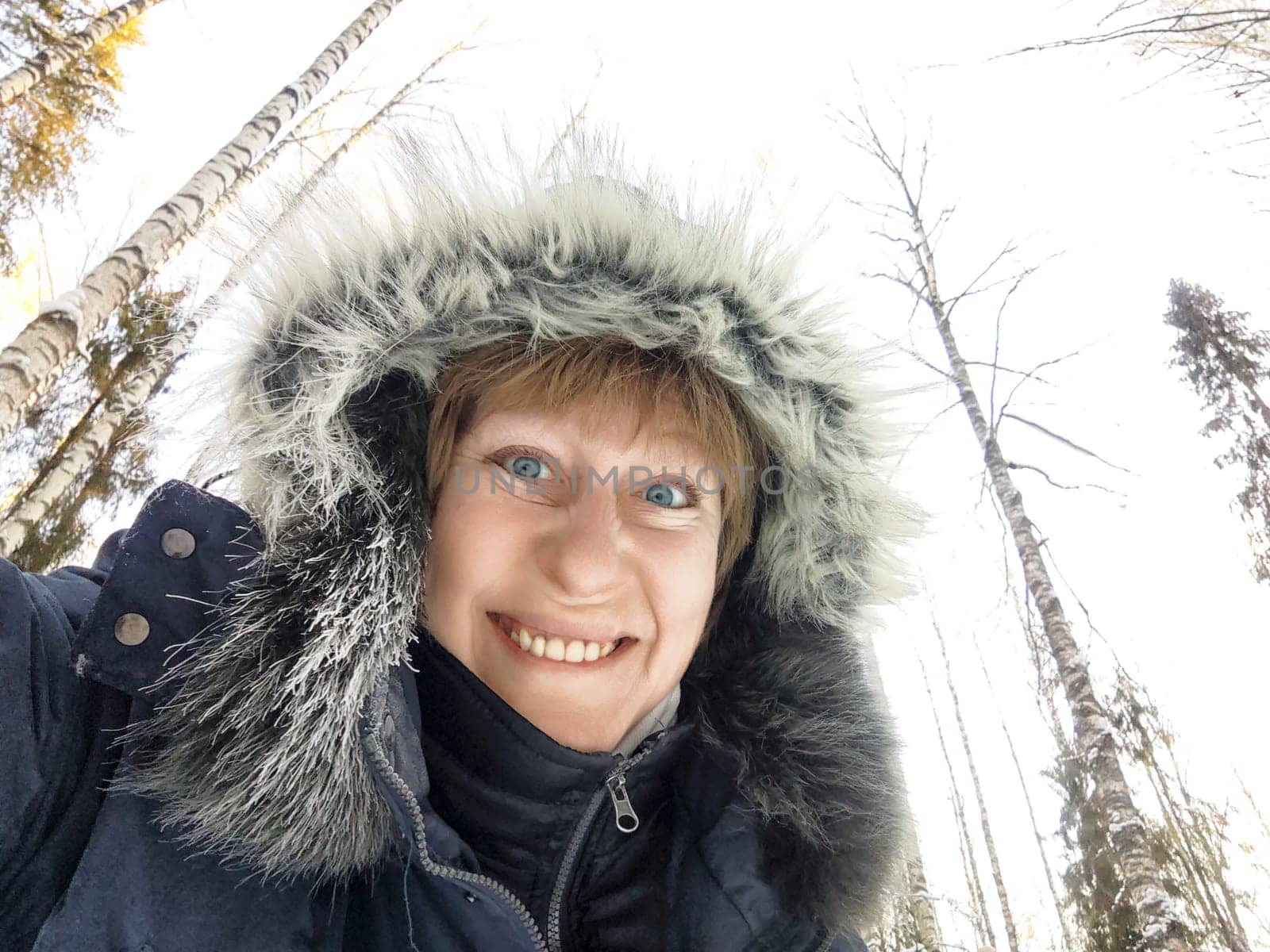 A woman enjoys a winter adventure while taking a cheerful selfie in a snowy forest, surrounded by tall trees.