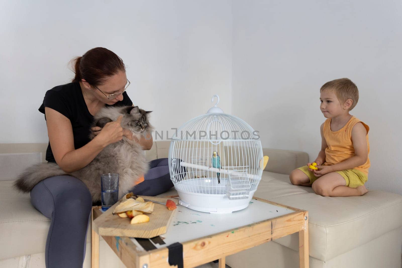Family enjoying time together with pets, mother cuddling cat while child looking at budgie in cage