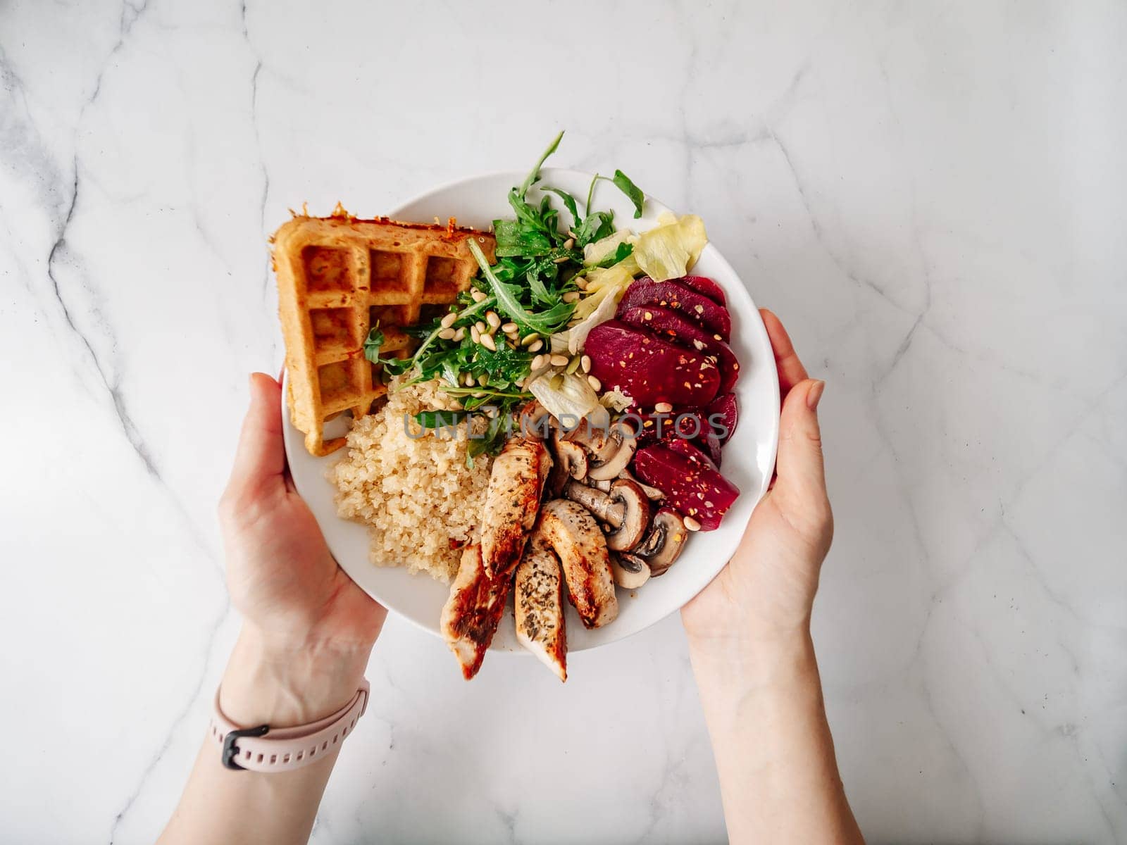 Top view of balanced gourmet dish in female hands over white marble tabletop. Bowl with chicken, mushrooms, beetroot, quinoa, green rucola, decorated pine nuts and carrot waffle. Healthy Food concept