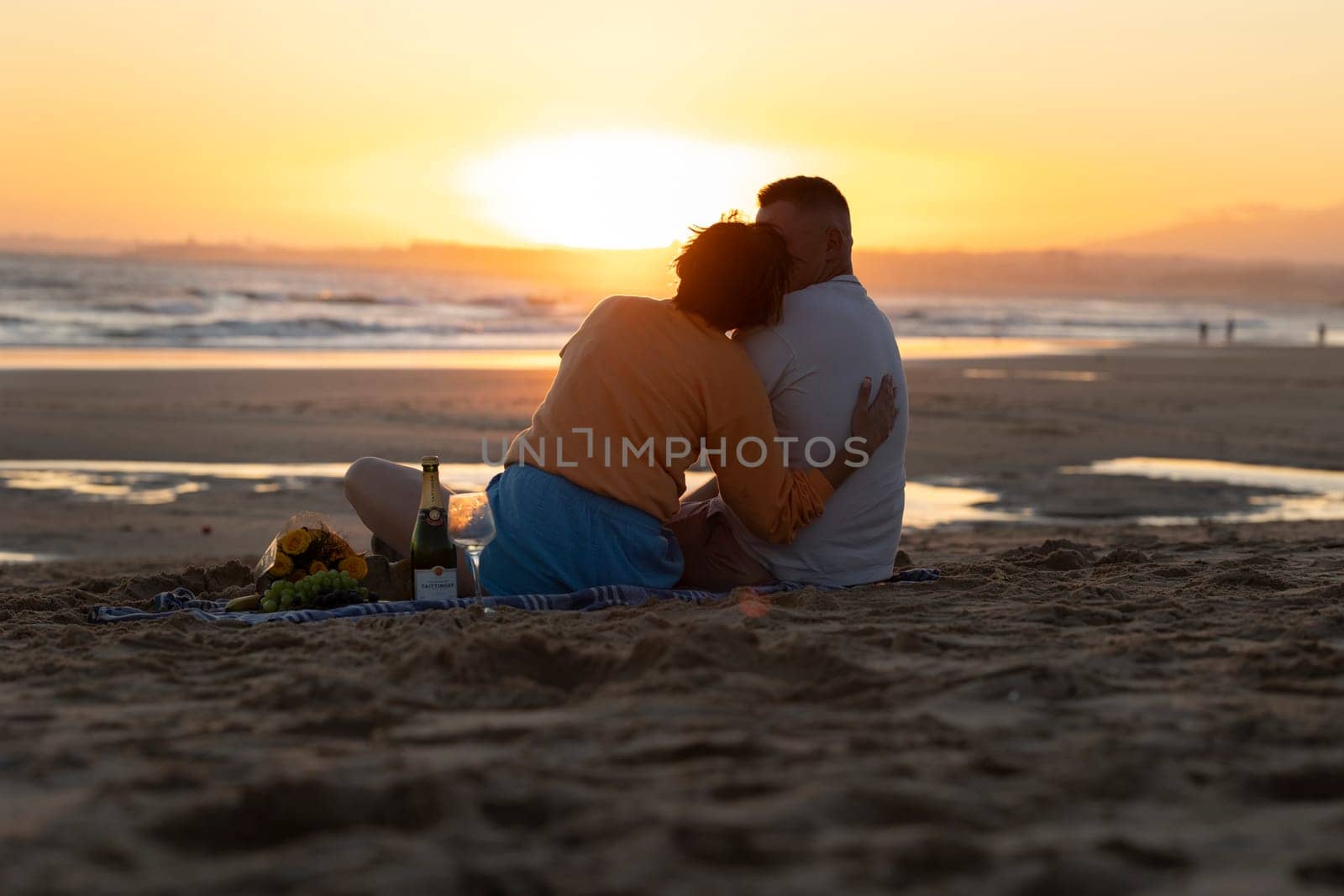 Couple embracing and enjoying a romantic sunset picnic on a sandy beach with sparkling wine and snacks