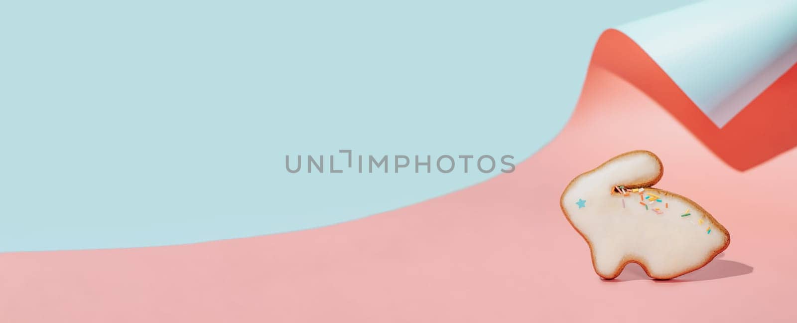 Easter bunny cookie on pink and blue curved paper pastel background with shadows. Happy Easter Day concept. Minimal Easter concept. Copy space for text. Easter cooking baking background.