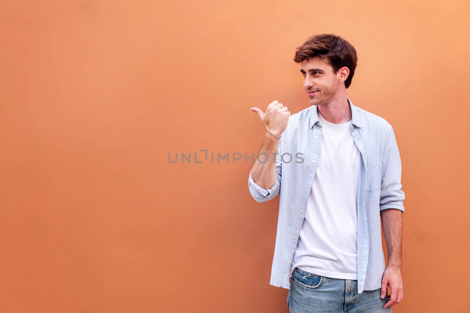 smiling young man in white shirt and blue jeans is pointing to the side with the thumb, copyspace for text