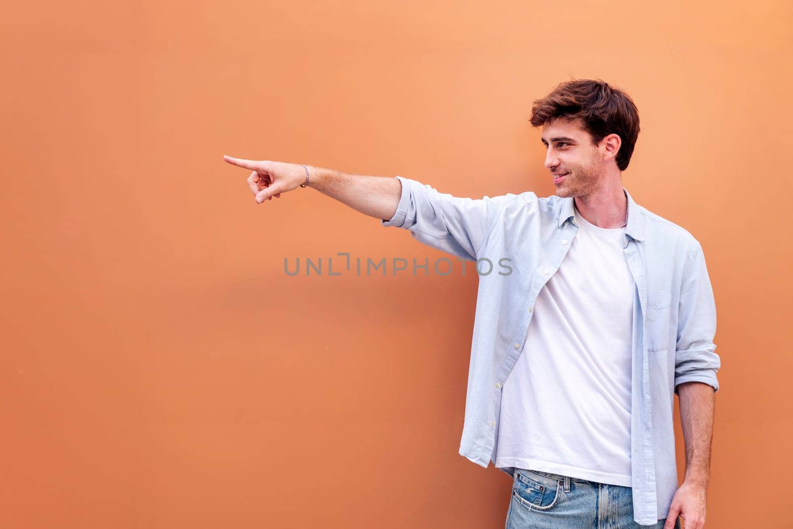 smiling young man in white shirt and blue jeans is pointing to the side, copyspace for text
