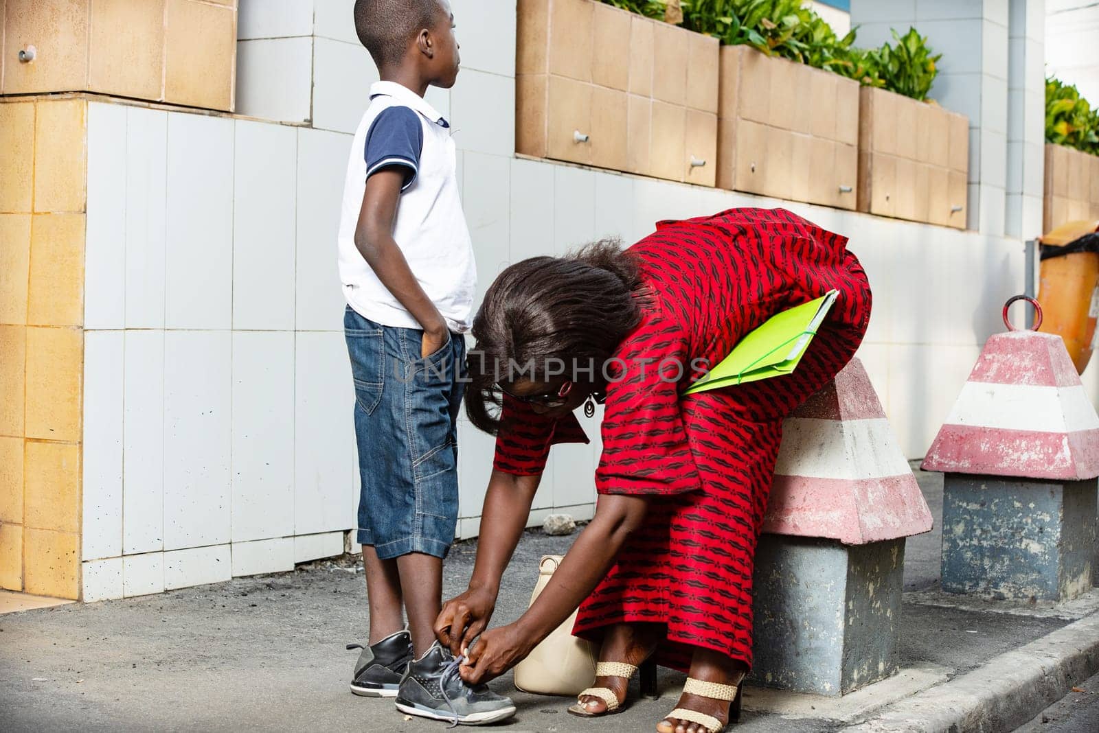Mature African woman in bent red loincloth, with her green document between her chest and her knees, is attaching the shoe laces of an arrested child in the street.