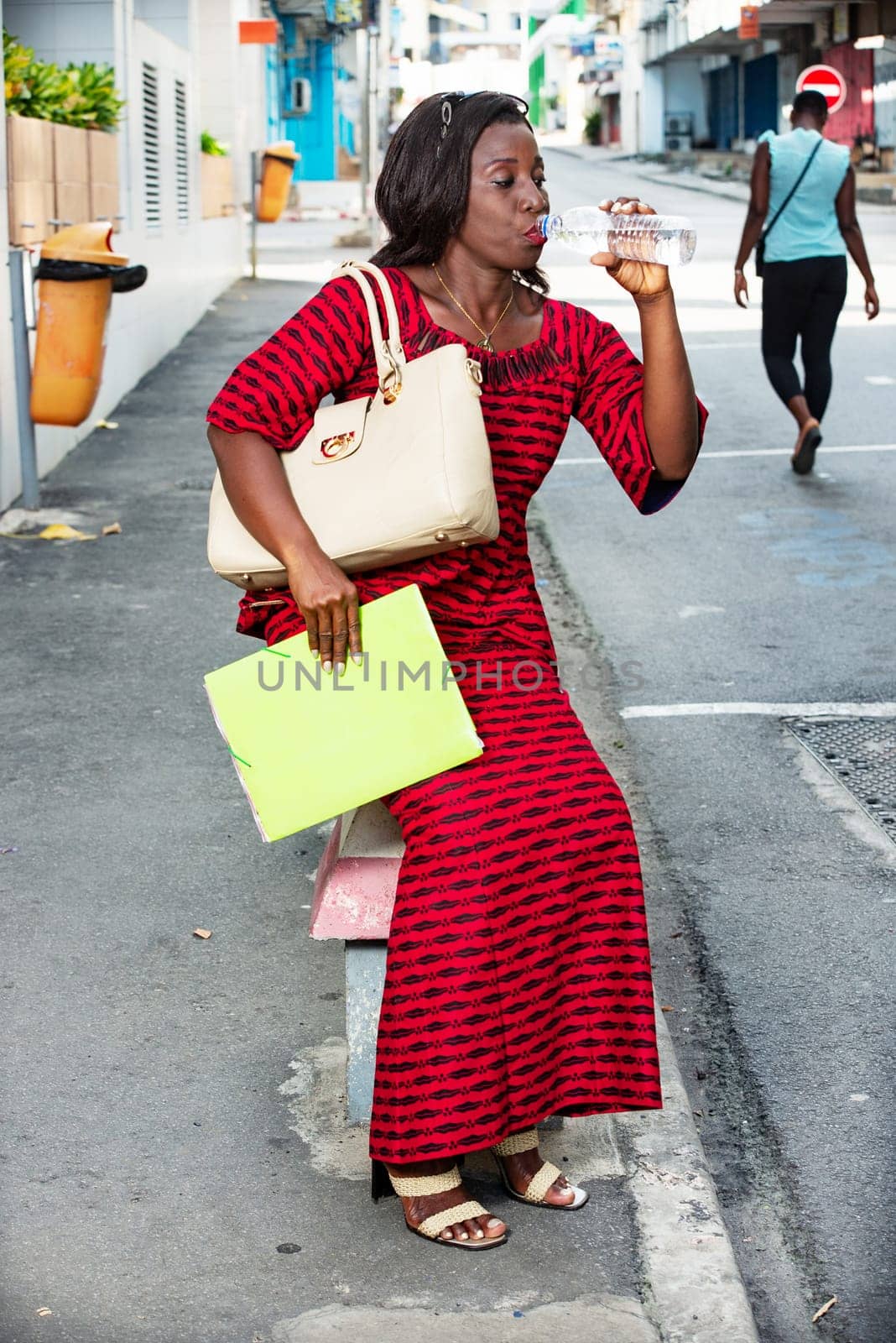 businesswoman in red dress holding a bag and documents, sitting on a concrete wall on the side of the road, off offices drinking bottled water