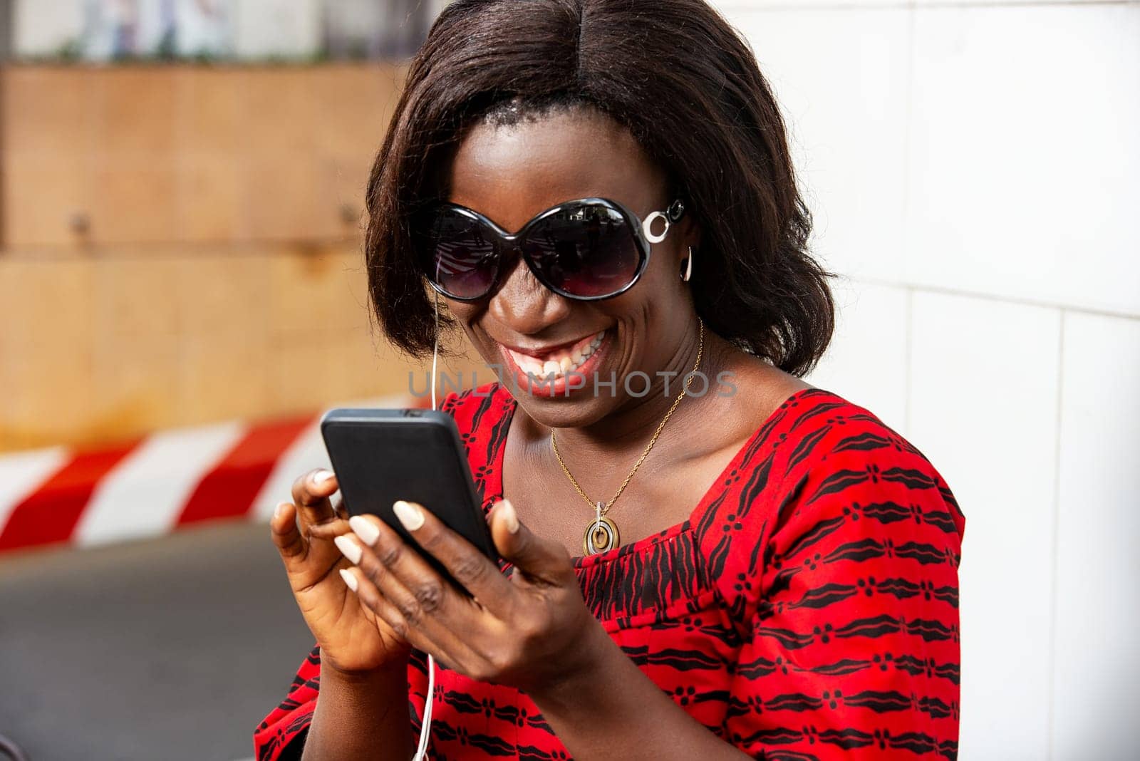 Happy woman in red outfit with sunglasses listening to music with headphones, outdoors during break while watching on a cell phone.