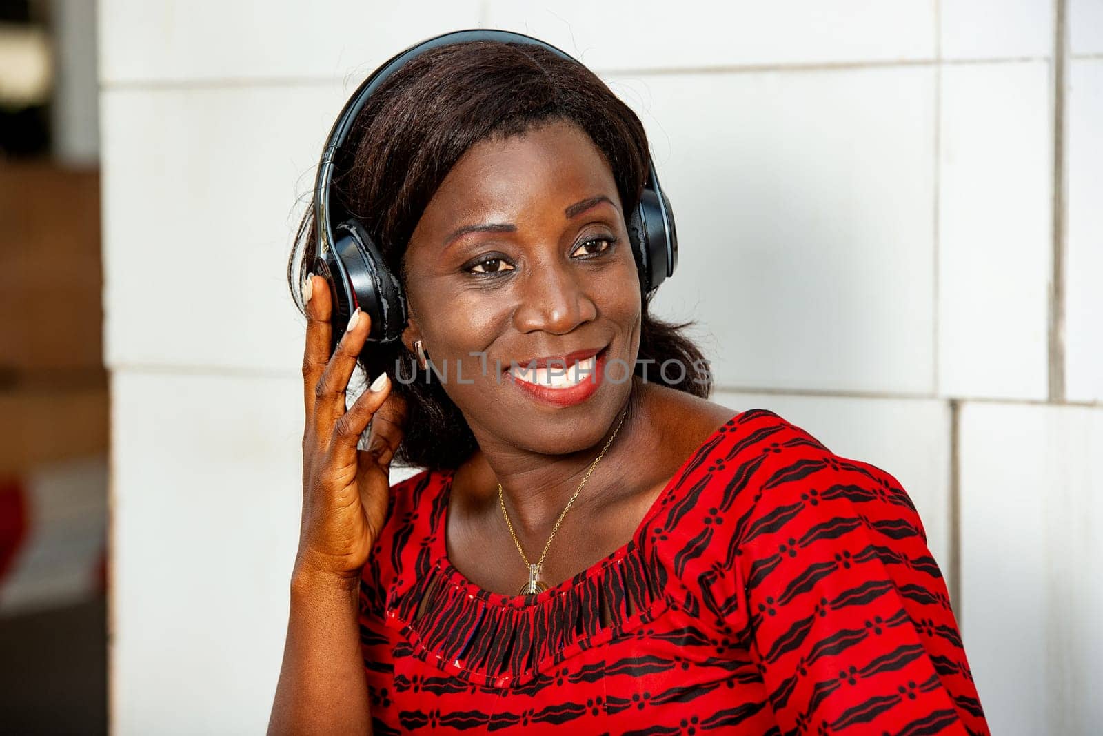 beautiful African adult woman under a beautiful red loincloth, is having fun listening to music in the street, with a headset earplug smiling