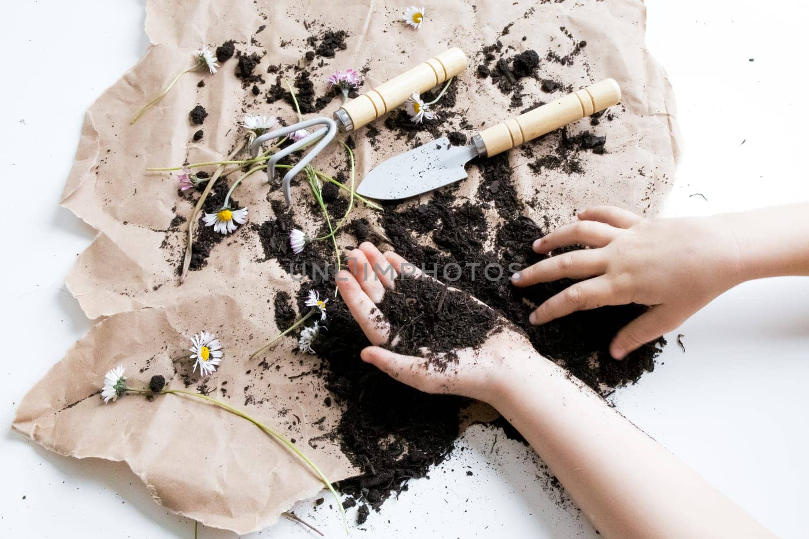 Childrens hands gardening with soil and tools on brown paper with flowers.