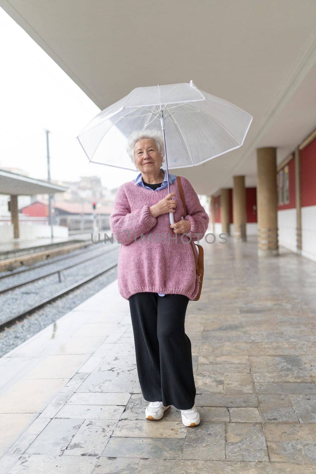 Elderly woman waiting for train arrival holding a transparent umbrella at rainy train station platform