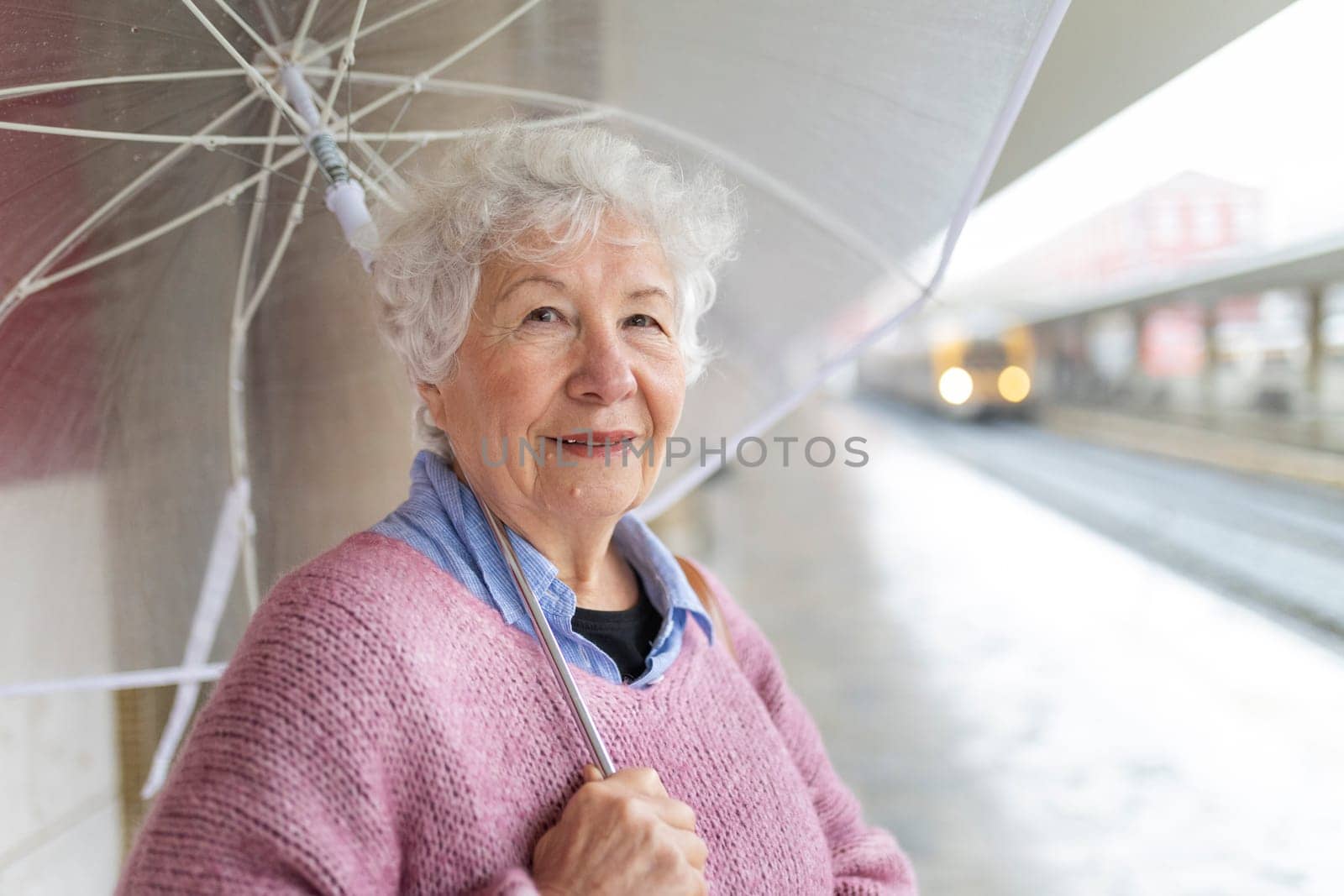 Senior woman smiling while holding a transparent umbrella at a train station on a rainy day