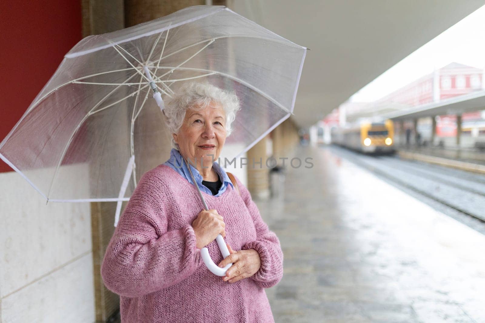 Elderly woman holding transparent umbrella waits at train station on a rainy day