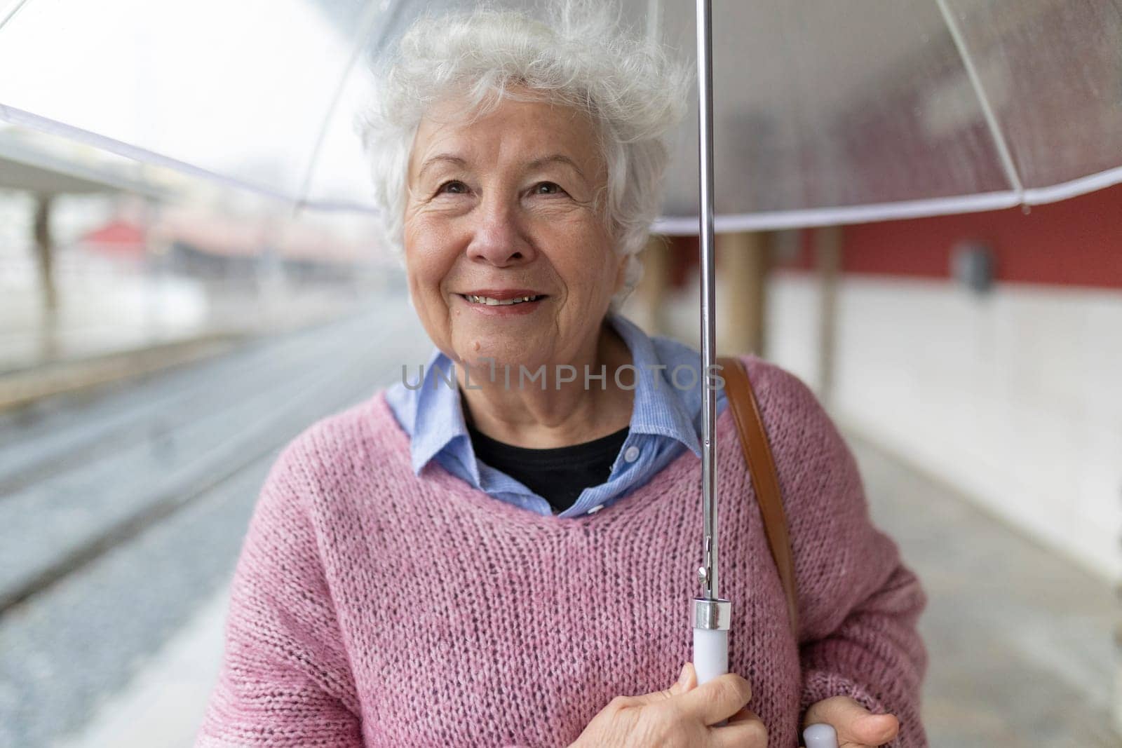 Happy elderly woman holding umbrella at train station platform on rainy day