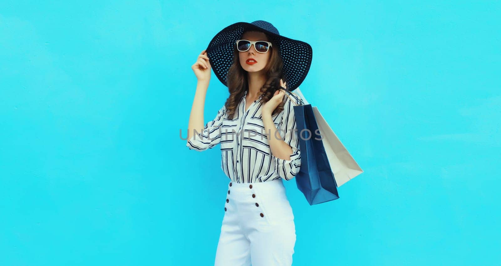 Stylish beautiful young woman posing with shopping bags in summer straw hat, white striped shirt looking away standing on blue wall background