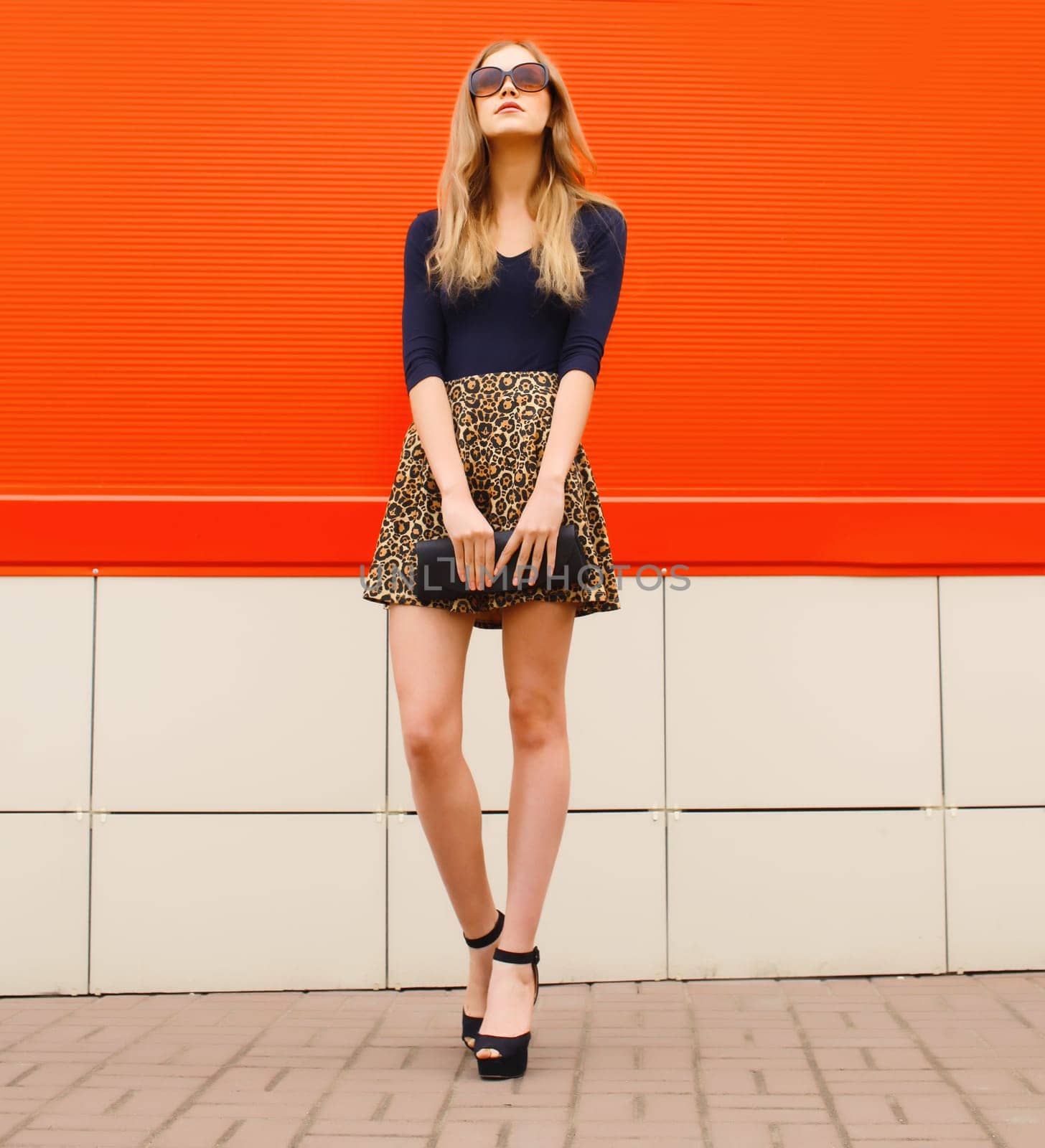 Beautiful stylish blonde young woman full length in leopard print skirt in heels with black handbag clutch standing on red wall, city street