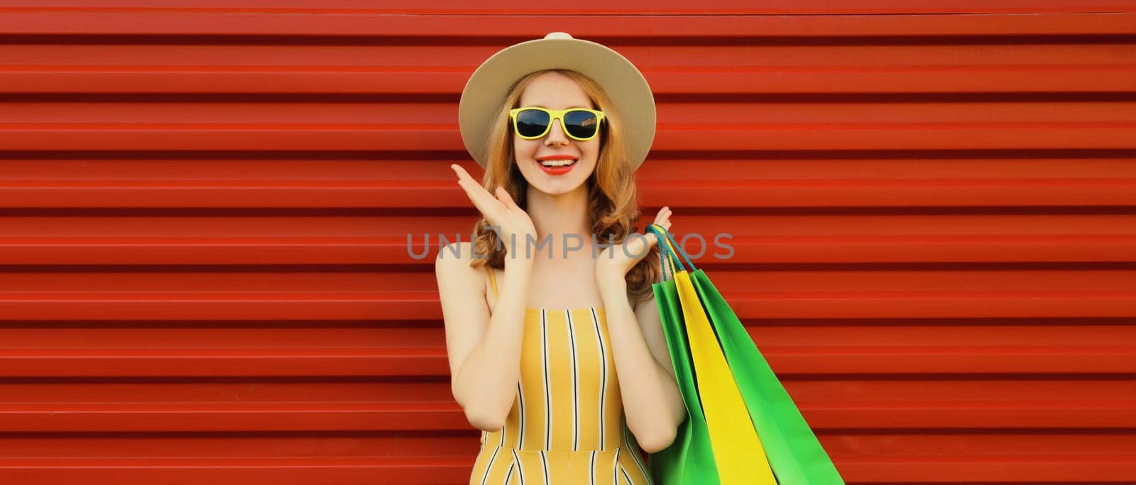 Fashion portrait, surprised happy beautiful young woman smiles posing with shopping bags in summer straw hat, looking at camera standing on red wall background
