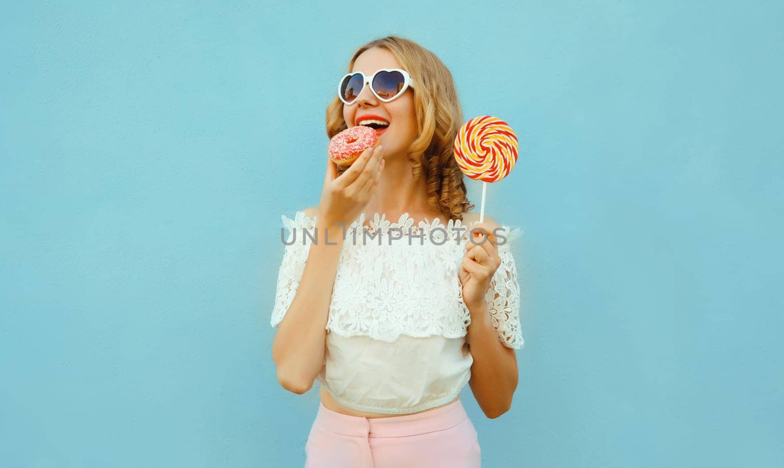Happy cheerful young woman eating sweet donut with lollipop having fun wearing a heart shaped sunglasses standing on blue background