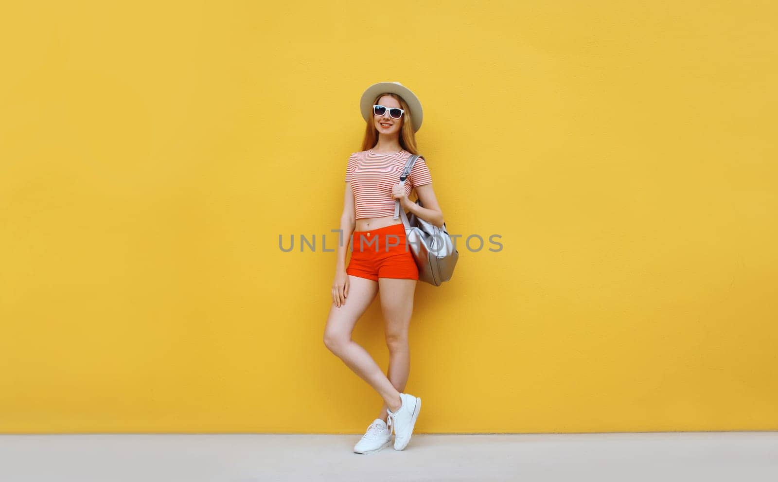 Beautiful young woman full length standing in summer straw hat posing with backpack on vivid yellow background