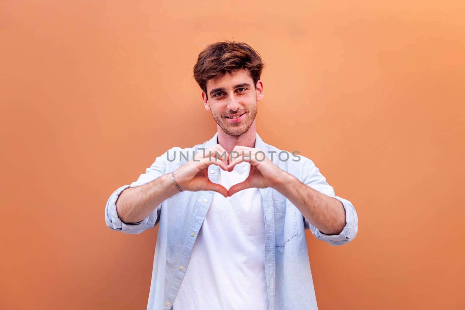young man standing in front of orange wall doing a heart shape with hands while looking at camera, he is wearing a blue shirt and white t-shirt, concept of love, copyspace for text