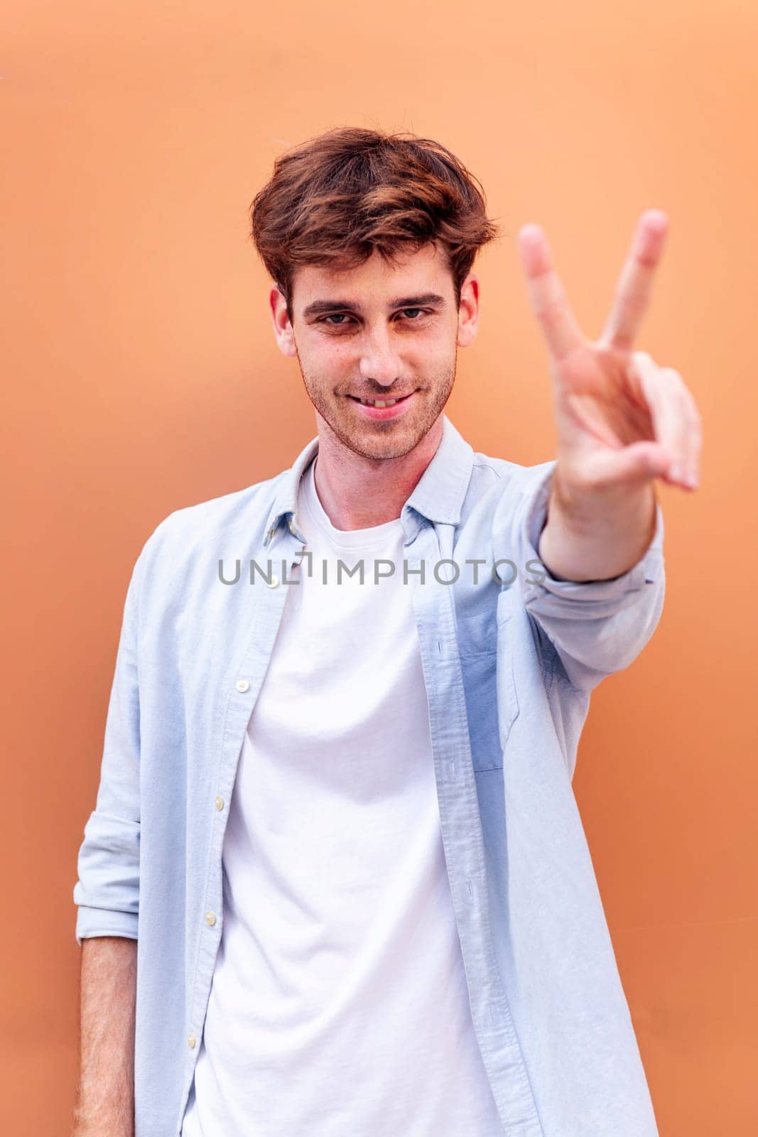 young man standing in front of orange wall holding up two fingers in victory sign and smiling looking at camera, concept of peace, love and positivity