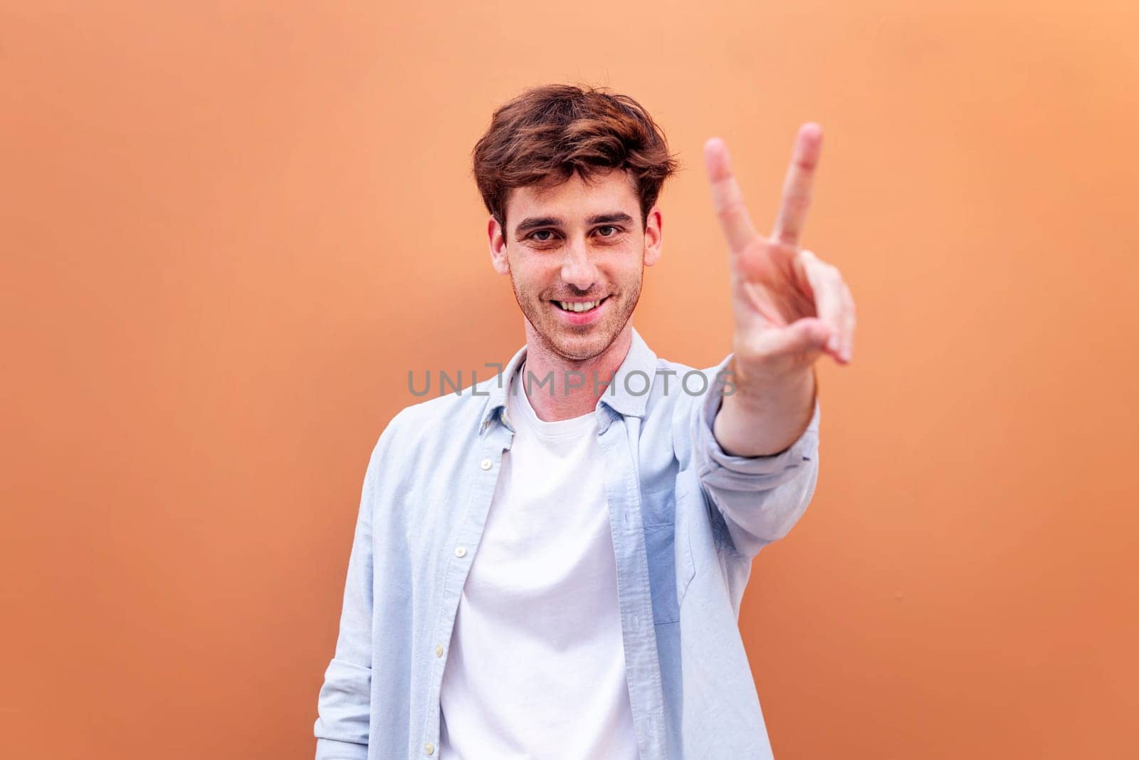 young man standing in front of orange wall holding up two fingers in victory sign and smiling looking at camera, concept of peace, love and positivity, copyspace for text