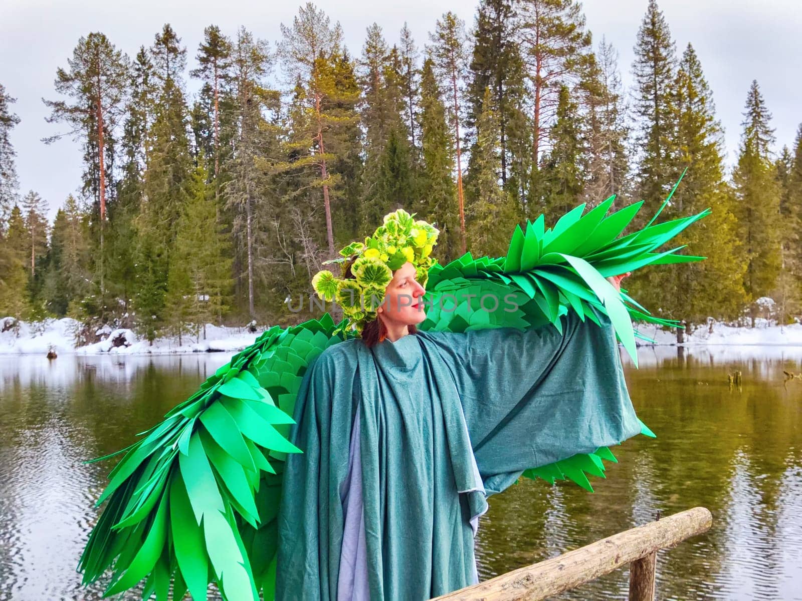 Girl wearing a green dress and wings stands by a tranquil lake, celebrating the arrival of spring and the enchanting spirit of nature.