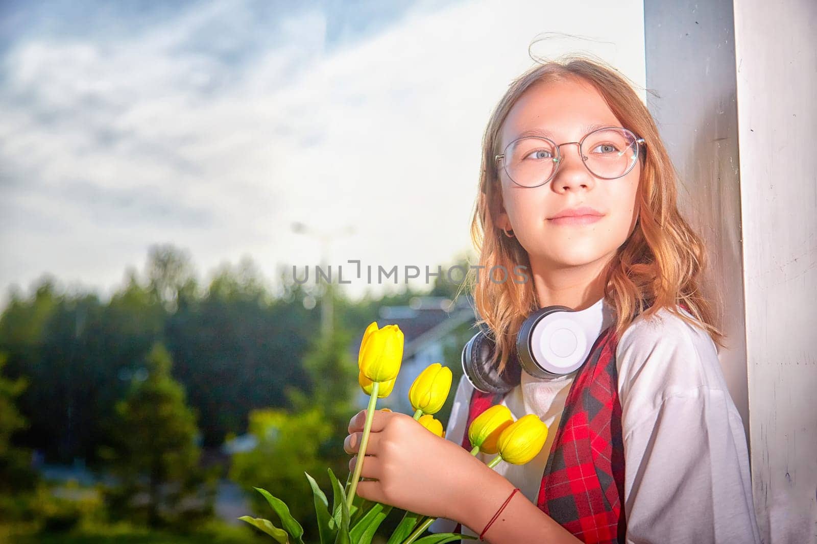 Girl holds vibrant yellow tulips while standing outside, enjoying the sunny weather and wearing headphones for music.
