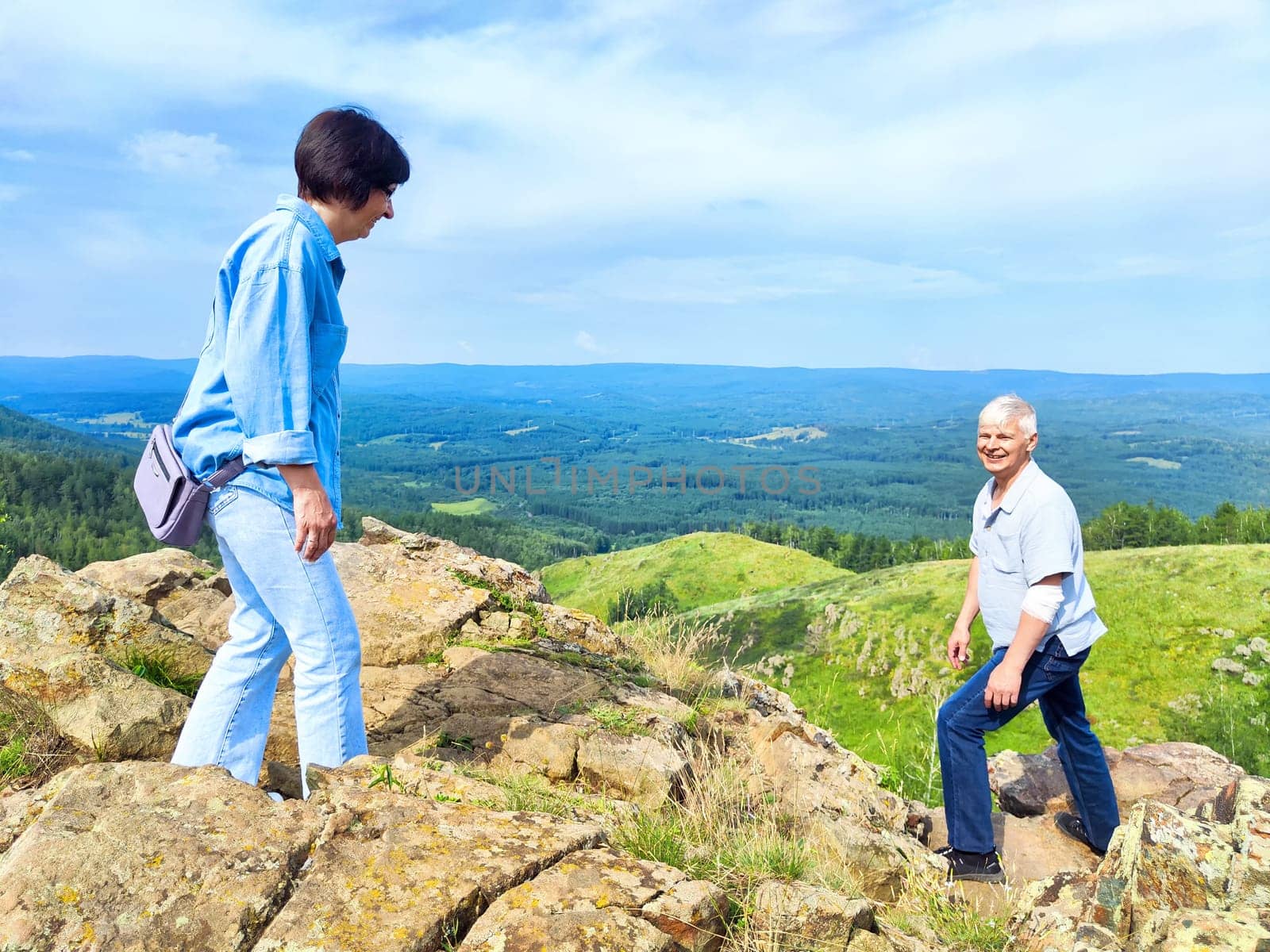 Two friends explore a rocky hilltop while surrounded by lush green valleys and a bright blue sky during summer.