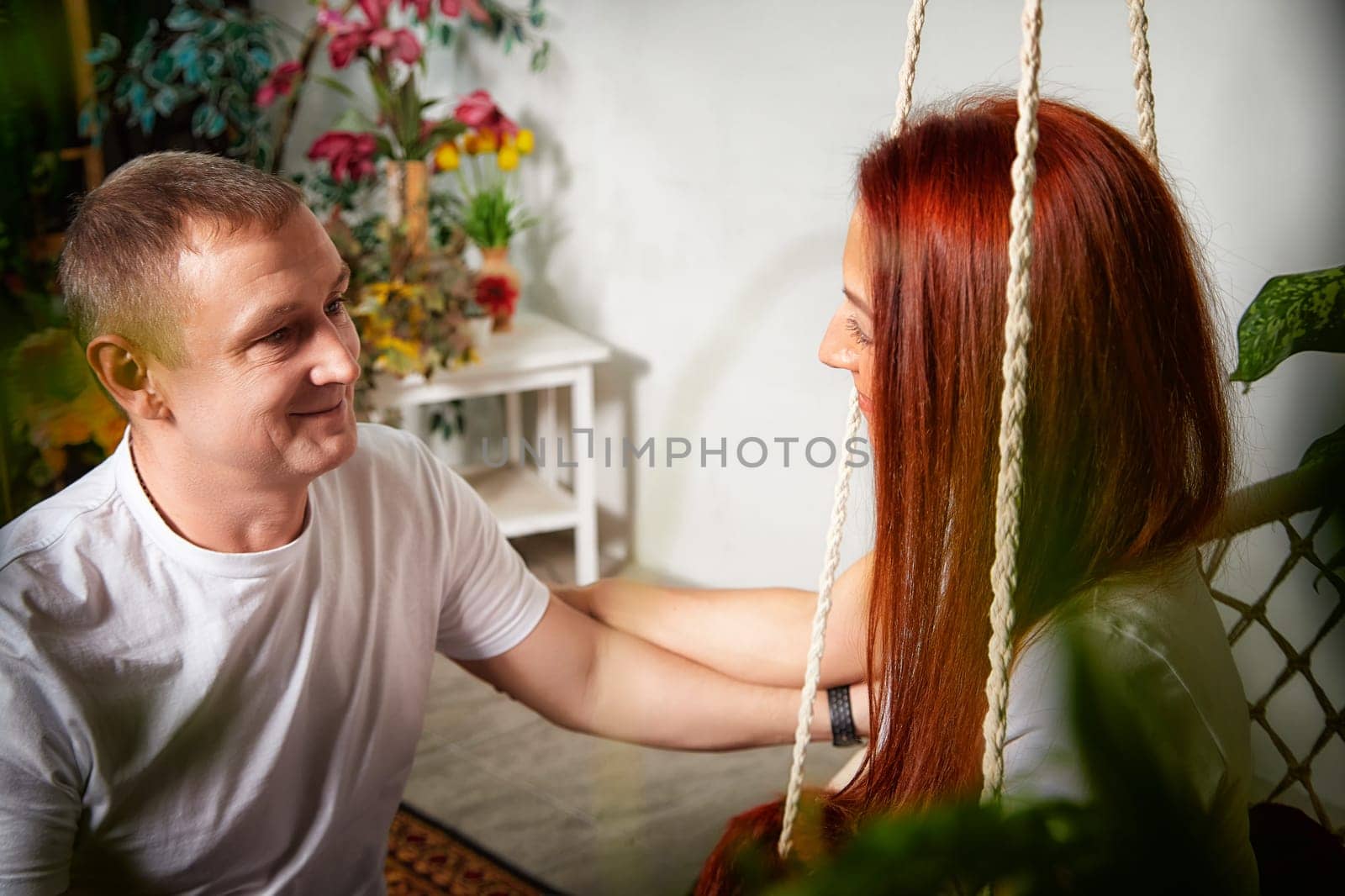 A woman with red hair and her husband enjoy a gentle exchange while sitting in a vibrant room surrounded by plants.