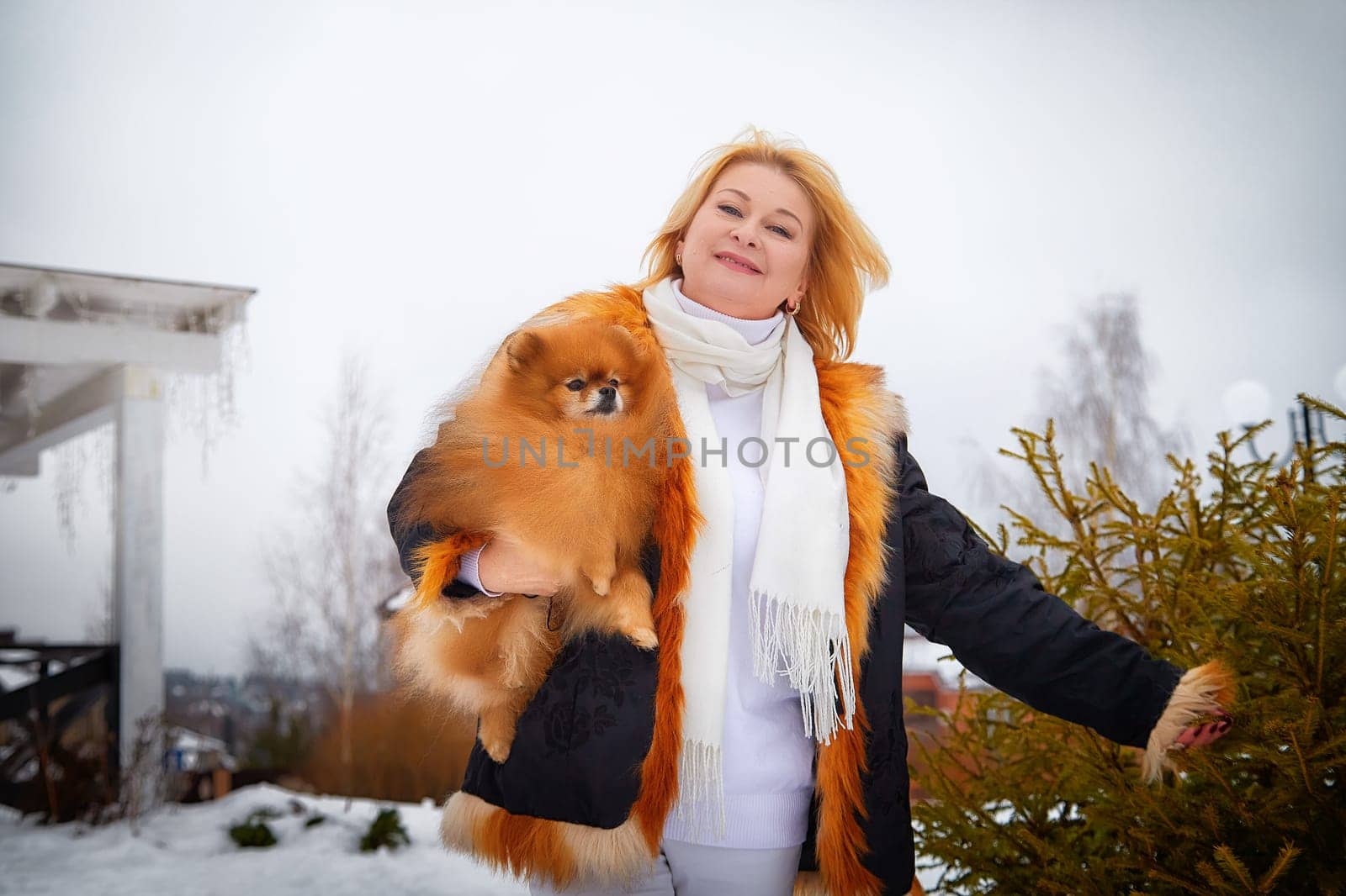 Blonde woman dressed warmly walks in the snow while holding her fluffy red Pomeranian close to her chest.