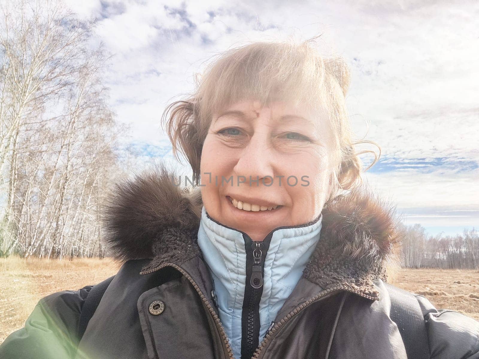 A woman smiles for a selfie while surrounded by trees under a partly cloudy sky on a clear day.