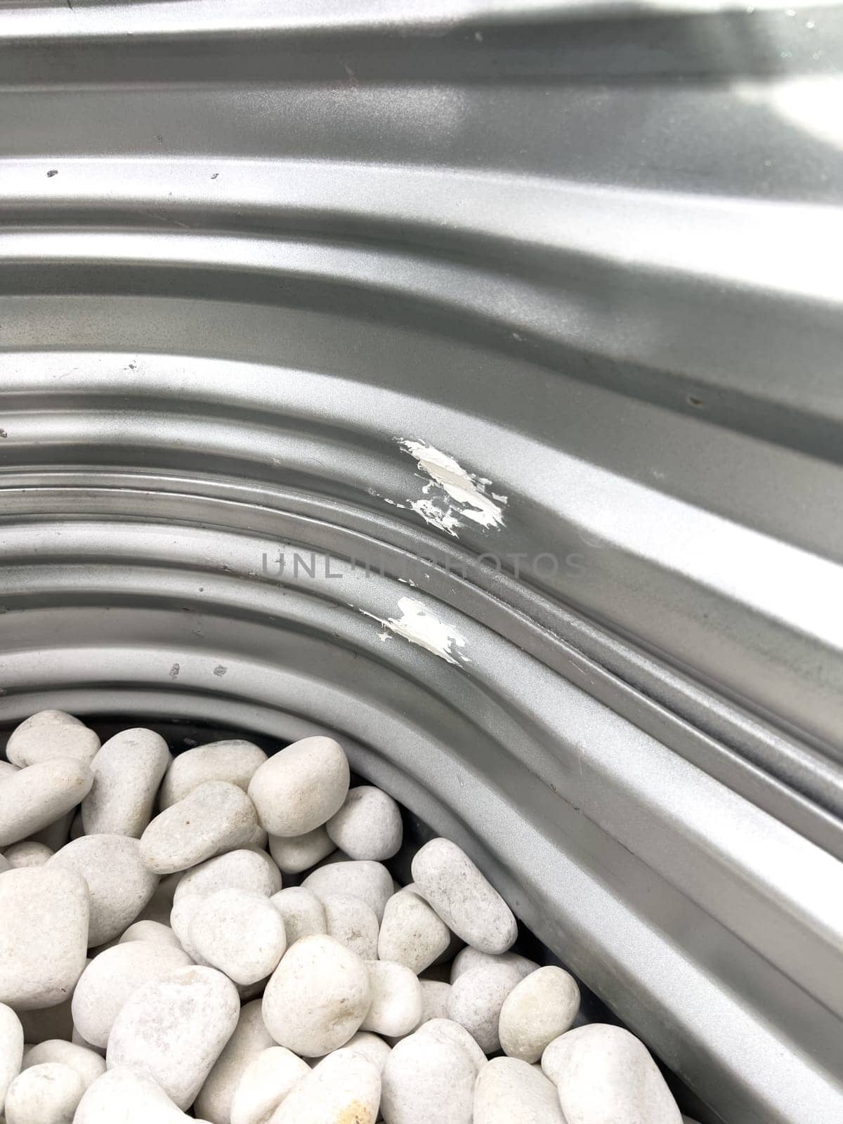 A close-up view of a corrugated metal window well with white decorative rocks at the base. The metal surface has minor scratches and wear, reflecting natural light. This functional design is commonly used for basement windows to allow light and ventilation while preventing soil erosion and water buildup.