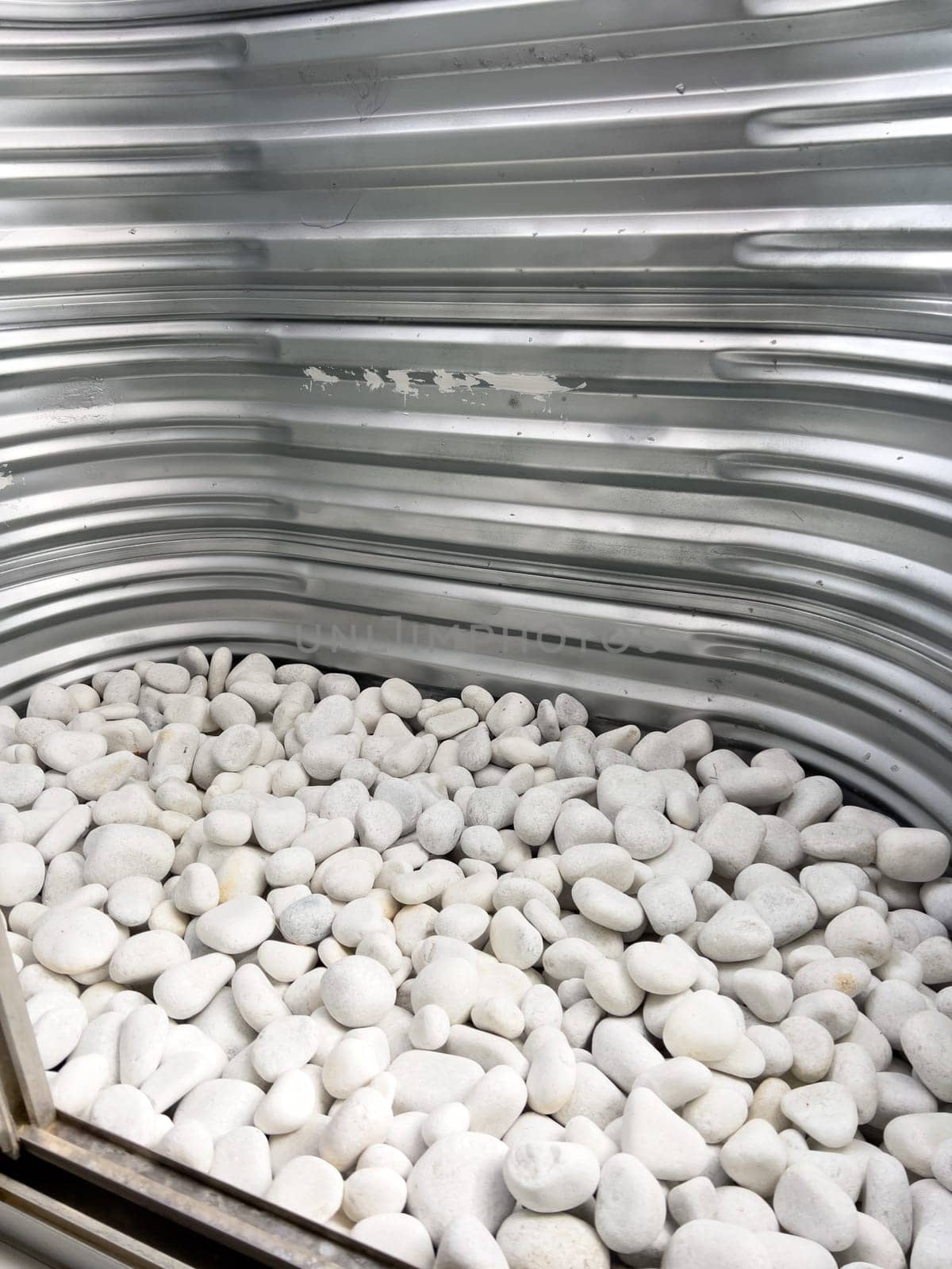 A close-up view of a corrugated metal window well with white decorative rocks at the base. The metal surface has minor scratches and wear, reflecting natural light. This functional design is commonly used for basement windows to allow light and ventilation while preventing soil erosion and water buildup.