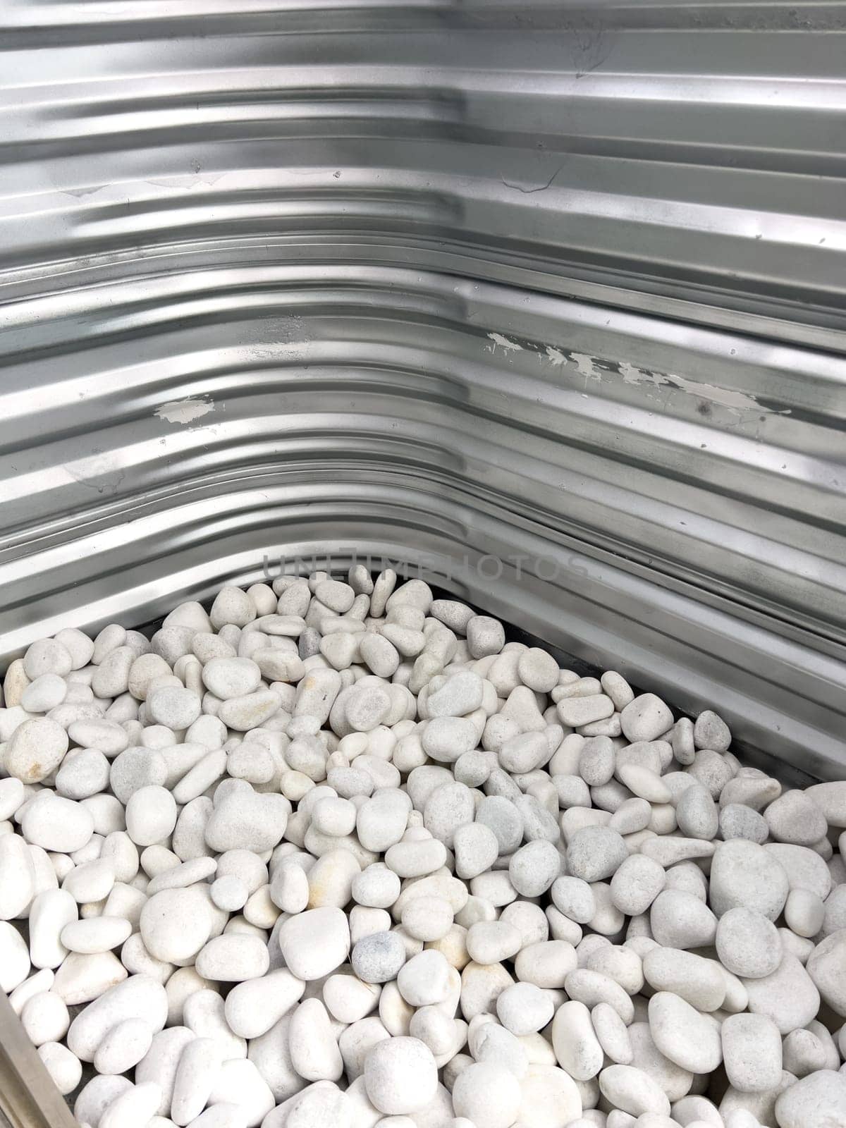 A close-up view of a corrugated metal window well with white decorative rocks at the base. The metal surface has minor scratches and wear, reflecting natural light. This functional design is commonly used for basement windows to allow light and ventilation while preventing soil erosion and water buildup.