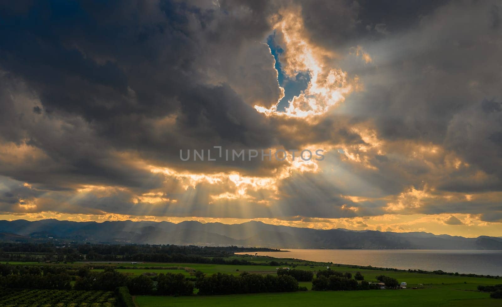 Golden beams of light break through dark clouds, illuminating a peaceful lake surrounded by lush greenery.