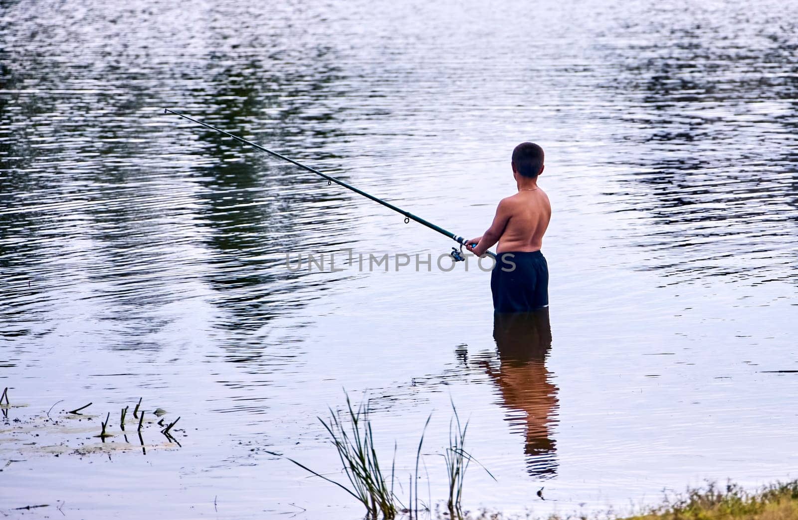 A young boy stands in a lake, holding a fishing rod and casting his line into the water. The sun reflects off the rippling surface.